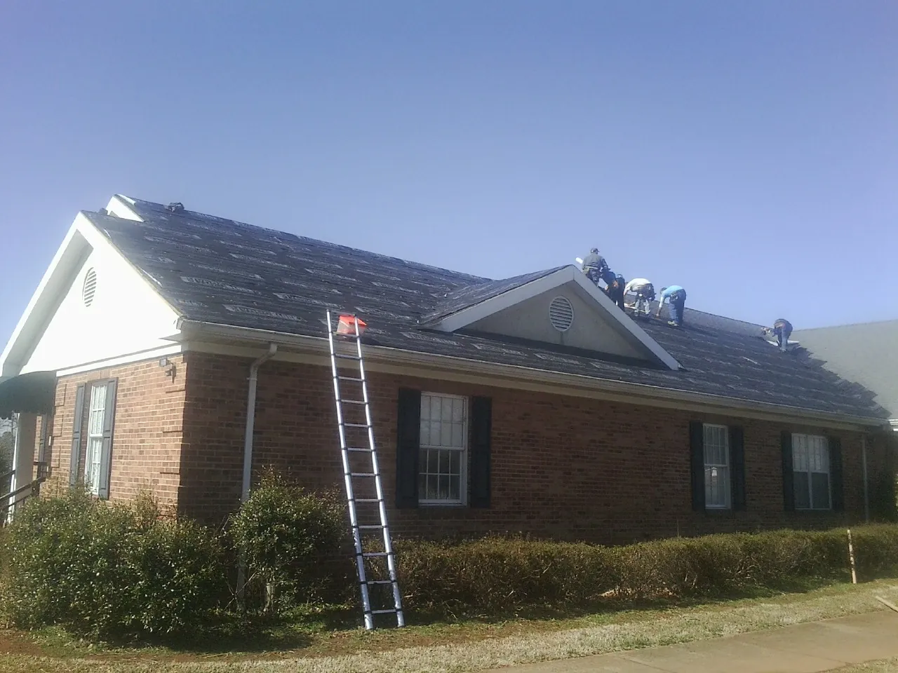 Crew tearing shingles off a brick ranch roof with a ladder set — Emergency Roof Repair work by our crew in Ashippun