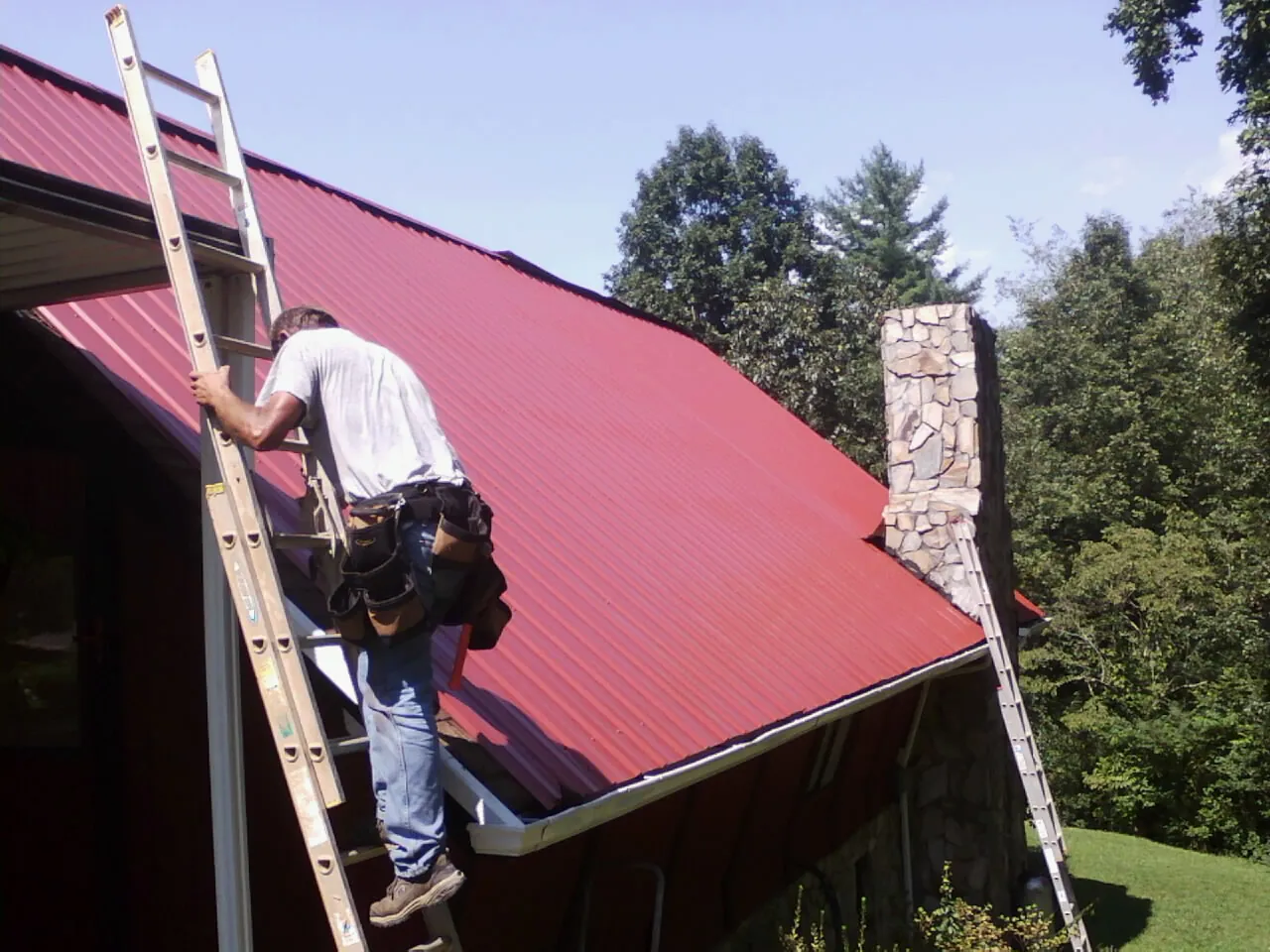 Roofer on a ladder beside a red metal roof and stone chimney — Soffit & Fascia Repair by Roofing Brothers in Ashippun